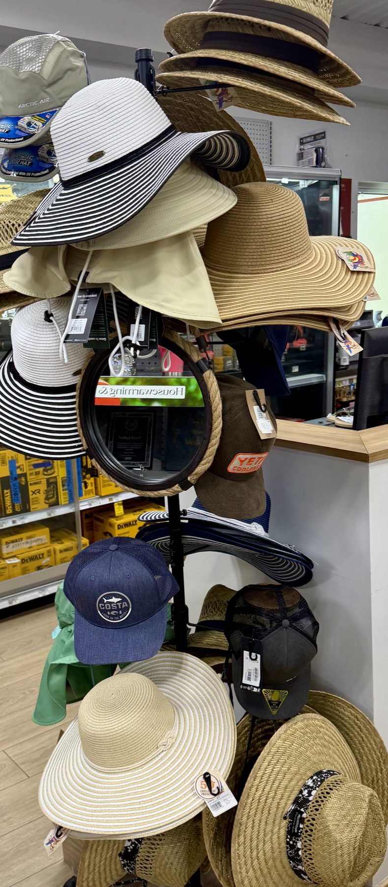 Display rack filled with wide-brim sun hats, straw hats, YETI caps, and Costa Del Mar hats at Hessler Paint & Hardware in Delray Beach, Florida.
